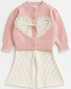 Conjunto infantil feminino com cardigan rosa de tricô com aplicação de coração rendado e calça off-white de malha canelada.