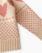 Detalhe da alça com botão do macacão infantil de tricô, com estampa de corações rosa sobre fundo bege.